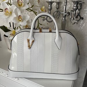 Puede incluir: Un bolso blanco con un acabado brillante y un dise&ntilde;o estructurado. El bolso presenta una mezcla de paneles lisos y texturizados, con un asa superior y detalles de herrajes dorados. El bolso tiene un ribete negro.