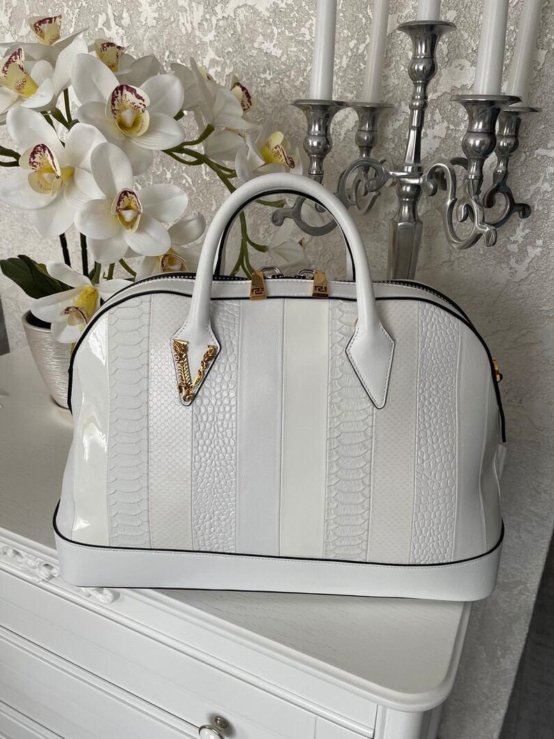 Puede incluir: Un bolso blanco con un acabado brillante y un dise&ntilde;o estructurado. El bolso presenta una mezcla de paneles lisos y texturizados, con un asa superior y detalles de herrajes dorados. El bolso tiene un ribete negro.