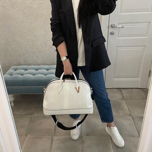 Puede incluir: Un bolso blanco con un patr&oacute;n texturizado y una correa negra es sostenido. El bolso tiene un detalle en forma de V de color dorado. La persona lleva una blazer negra, una camiseta blanca, vaqueros azules y zapatillas blancas.