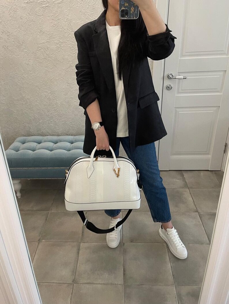 Puede incluir: Un bolso blanco con un patr&oacute;n texturizado y una correa negra es sostenido. El bolso tiene un detalle en forma de V de color dorado. La persona lleva una blazer negra, una camiseta blanca, vaqueros azules y zapatillas blancas.