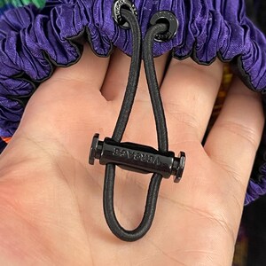 May include: A purple pleated fabric garment featuring a black elastic drawstring cord with a black plastic toggle. The toggle is engraved with the word VERSACE, and the cord passes through two black metal eyelets also engraved with the brand name.