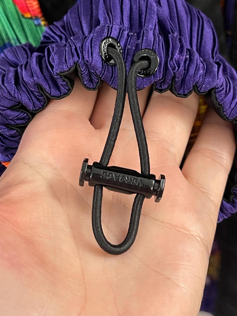 May include: A purple pleated fabric garment featuring a black elastic drawstring cord with a black plastic toggle. The toggle is engraved with the word VERSACE, and the cord passes through two black metal eyelets also engraved with the brand name.