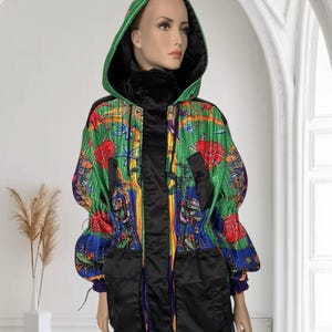 May include: A hooded jacket featuring a vibrant, multi-colored graphic print with green, blue, and red tones. The garment includes a black front placket, black oversized pockets, and a black inner lining. The fabric has a pleated texture throughout.
