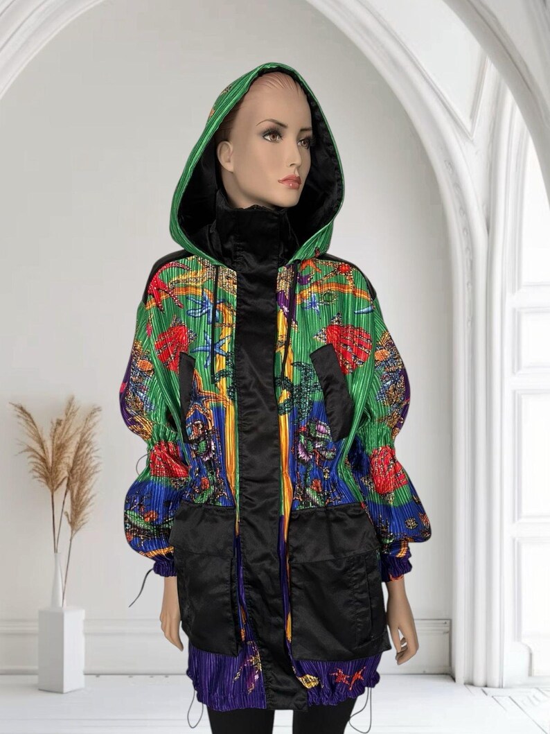 May include: A hooded jacket featuring a vibrant, multi-colored graphic print with green, blue, and red tones. The garment includes a black front placket, black oversized pockets, and a black inner lining. The fabric has a pleated texture throughout.