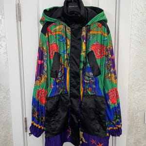 May include: A hooded jacket featuring a vibrant, pleated fabric with a colorful marine-themed print of seashells, starfish, and coral in shades of green, blue, yellow, and red. The jacket includes solid black panels, a front zipper, and pockets.