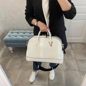 Puede incluir: Bolso de mano blanco con un patr&oacute;n texturizado y rayas verticales. El bolso tiene una asa superior y una correa negra para el hombro. Un logotipo dorado es visible en la parte delantera. El bolso lo sostiene una persona que lleva una blazer negra, una camiseta blanca y vaqueros azules.