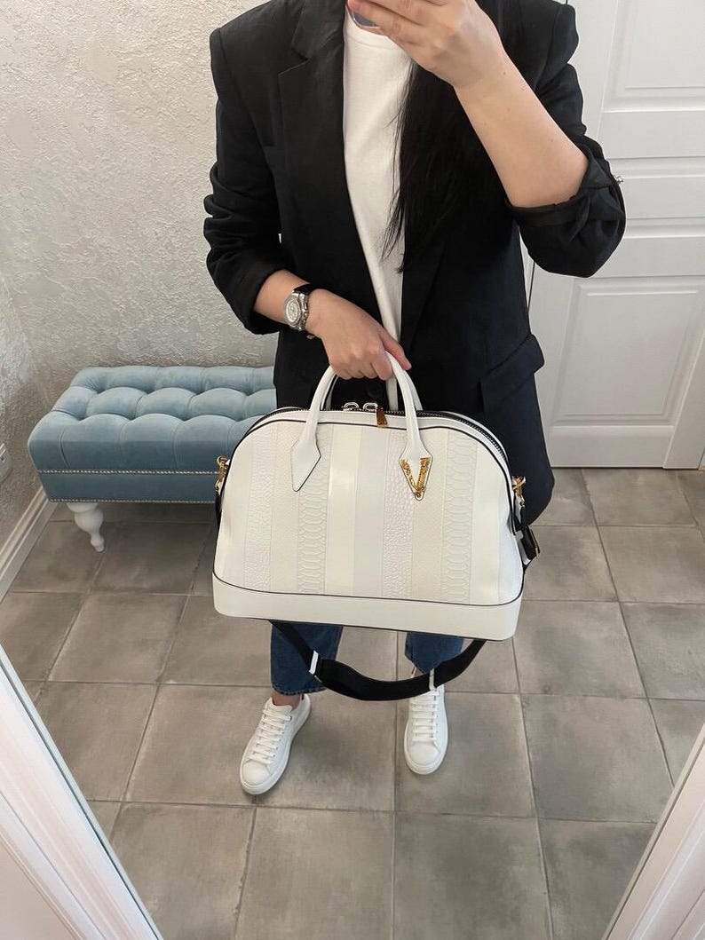 Puede incluir: Bolso de mano blanco con un patr&oacute;n texturizado y rayas verticales. El bolso tiene una asa superior y una correa negra para el hombro. Un logotipo dorado es visible en la parte delantera. El bolso lo sostiene una persona que lleva una blazer negra, una camiseta blanca y vaqueros azules.