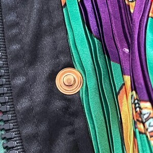 May include: A garment featuring a black fabric panel with a vertical zipper and a circular gold-toned metal snap button. Adjacent to the black section is a vibrant, pleated fabric panel displaying a colorful pattern of green, purple, and orange.