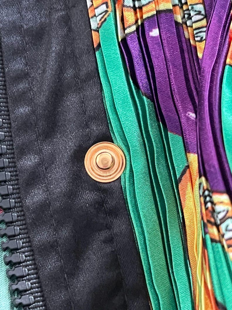 May include: A garment featuring a black fabric panel with a vertical zipper and a circular gold-toned metal snap button. Adjacent to the black section is a vibrant, pleated fabric panel displaying a colorful pattern of green, purple, and orange.