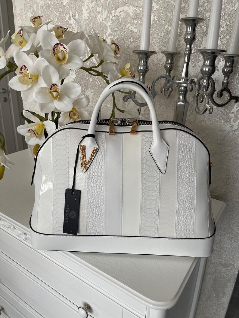 Puede incluir: Bolso de mano de cuero blanco con dise&ntilde;o a rayas. El bolso tiene una asa superior, cierre de cremallera y herrajes dorados. Una etiqueta negra con el logo de Versace cuelga del bolso.