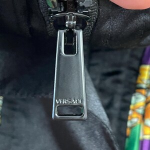 May include: A dark metallic zipper pull featuring the word VERSACE embossed in capital letters. The rectangular pull is attached to a black fabric garment with a visible portion of a colorful patterned fabric in green, yellow, and purple.
