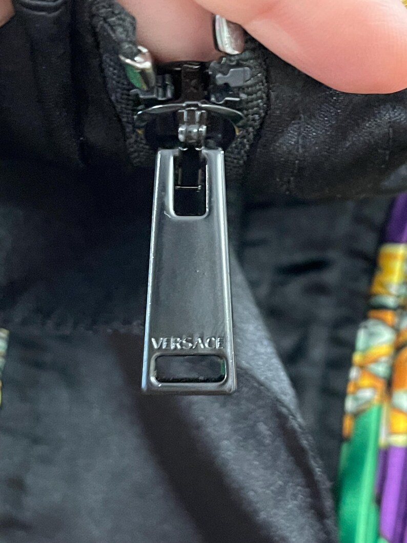 May include: A dark metallic zipper pull featuring the word VERSACE embossed in capital letters. The rectangular pull is attached to a black fabric garment with a visible portion of a colorful patterned fabric in green, yellow, and purple.