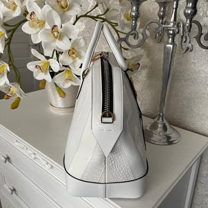 Puede incluir: Un bolso blanco con un acabado brillante, con un patr&oacute;n texturizado y una cremallera negra. El bolso tiene una forma estructurada con un asa superior y un tirador dorado. El bolso est&aacute; sobre una superficie blanca.