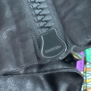 May include: A black garment featuring a heavy-duty zipper with a large, dark metal pull tab. The word VERSACE is embossed in capital letters on the surface of the zipper pull. The fabric appears to have a matte, textured finish with visible stitching.