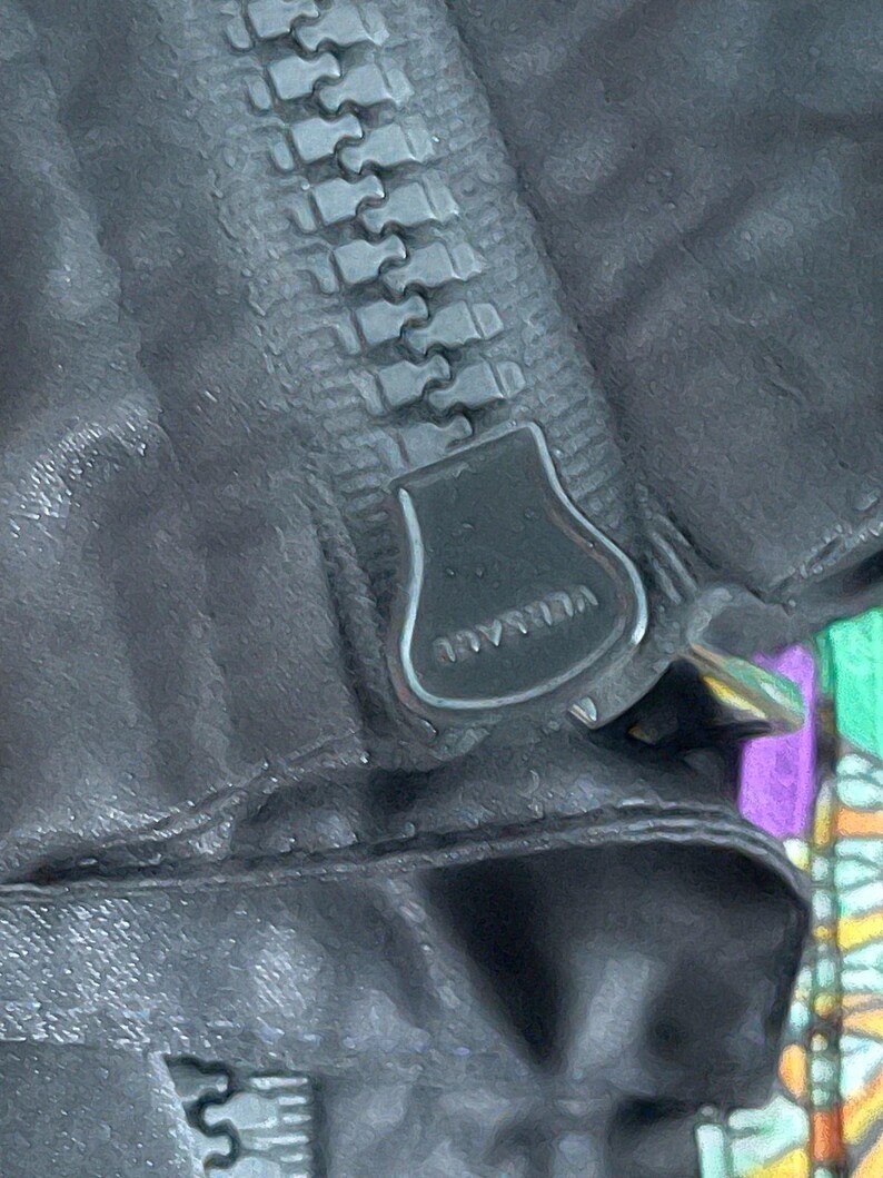 May include: A black garment featuring a heavy-duty zipper with a large, dark metal pull tab. The word VERSACE is embossed in capital letters on the surface of the zipper pull. The fabric appears to have a matte, textured finish with visible stitching.