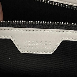 Puede incluir: Primer plano de una etiqueta de cuero blanco con la palabra "VERSACE" en relieve y el texto "MADE IN ITALY". La etiqueta est&aacute; cosida sobre una tela negra, con una cremallera plateada en la parte superior.
