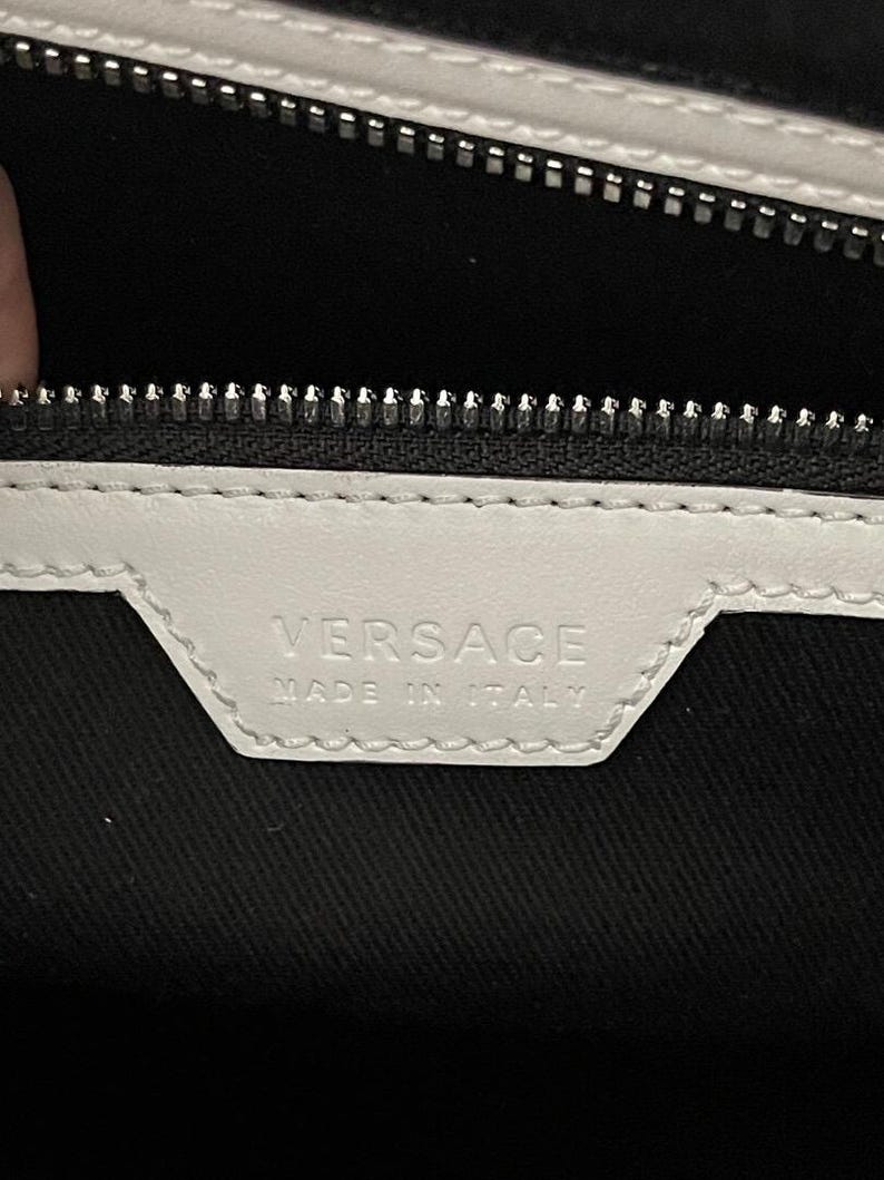 Puede incluir: Primer plano de una etiqueta de cuero blanco con la palabra "VERSACE" en relieve y el texto "MADE IN ITALY". La etiqueta est&aacute; cosida sobre una tela negra, con una cremallera plateada en la parte superior.