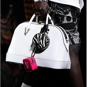 Puede incluir: Bolso de mano de cuero blanco con un patr&oacute;n texturizado, cremallera negra y herrajes dorados. Un peque&ntilde;o bolso redondo con estampado de cebra y un mini bolso rosa est&aacute;n adjuntos. Accesorio de moda.