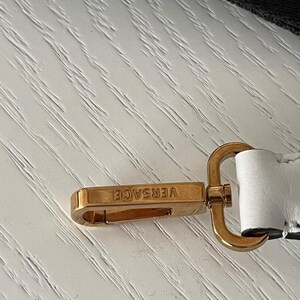 Puede incluir: Cierre met&aacute;lico dorado con el nombre de la marca "VERSACE" grabado. El cierre es rectangular con bordes redondeados y un lazo de conexi&oacute;n. El fondo es una superficie clara y texturizada.