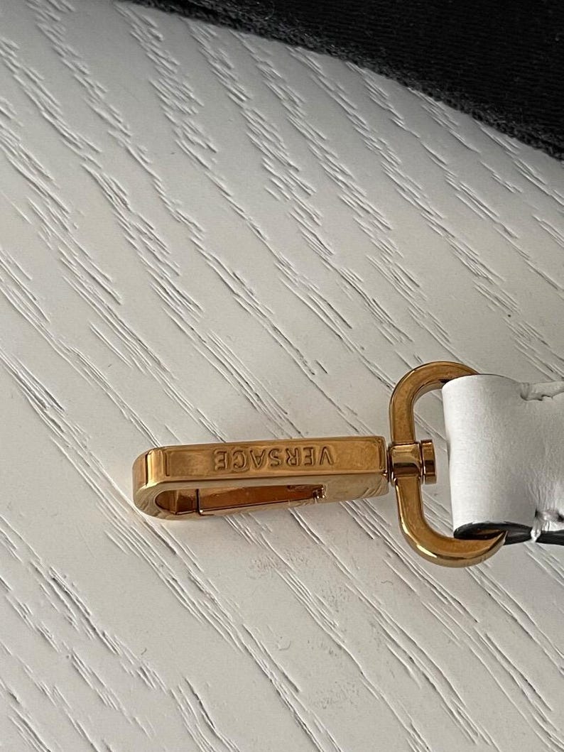 Puede incluir: Cierre met&aacute;lico dorado con el nombre de la marca "VERSACE" grabado. El cierre es rectangular con bordes redondeados y un lazo de conexi&oacute;n. El fondo es una superficie clara y texturizada.