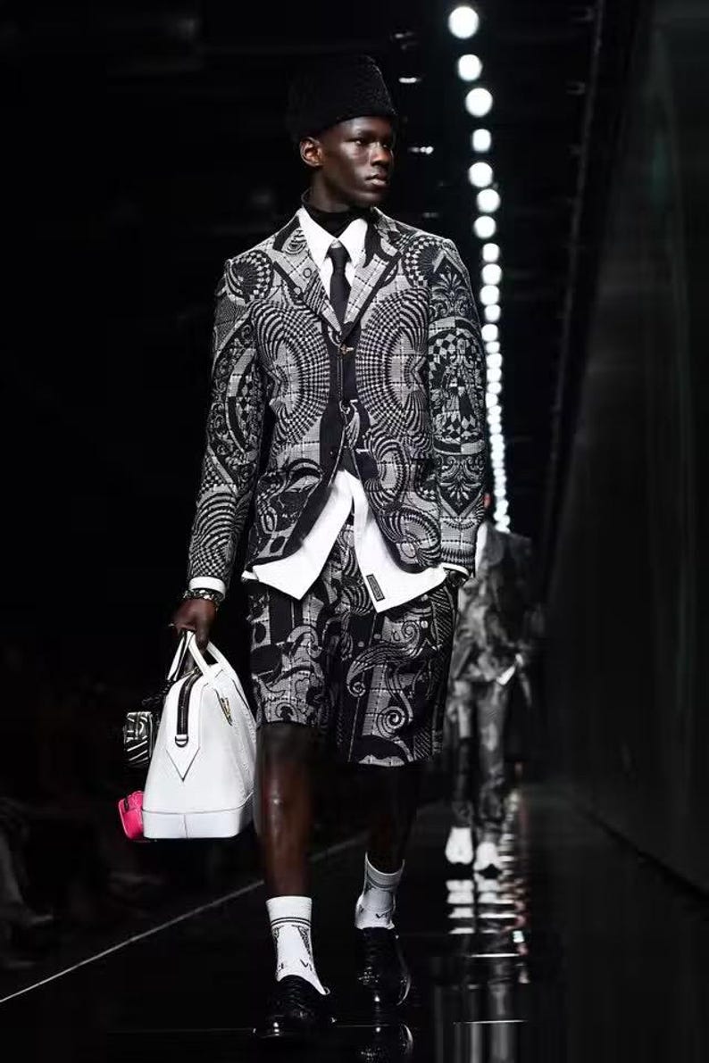 Puede incluir: Un traje con estampado en blanco y negro, compuesto por una americana y un pantal&oacute;n corto, se lleva con una camisa blanca y una corbata negra. El conjunto se completa con zapatos negros y calcetines blancos. Se lleva un bolso de mano blanco con detalles en negro.
