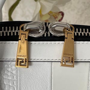 Puede incluir: Primer plano de un bolso de mano de cuero blanco con tiradores de cremallera dorados. Los tiradores de la cremallera son rectangulares con el nombre de la marca "VERSACE" y un dise&ntilde;o de greca. El bolso tiene una cremallera negra y un dise&ntilde;o a rayas blancas y negras.