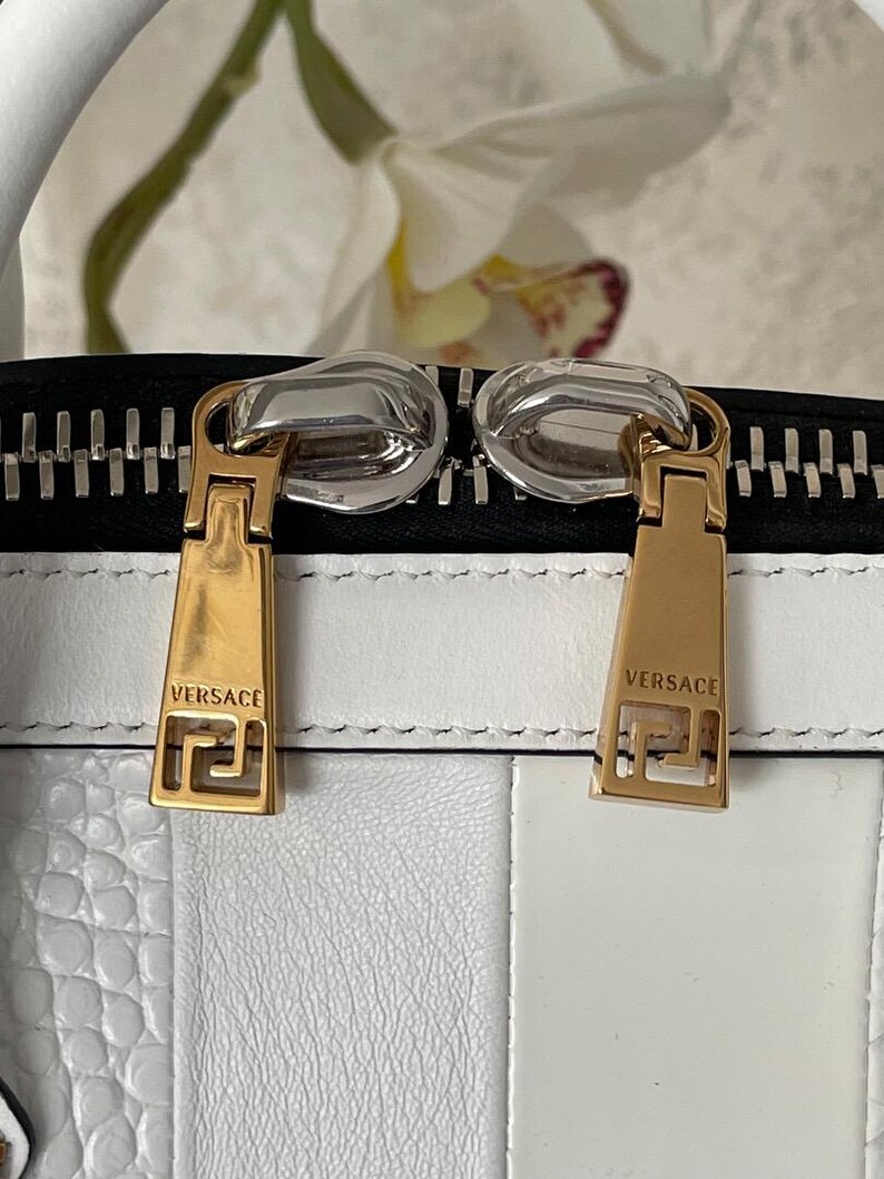 Puede incluir: Primer plano de un bolso de mano de cuero blanco con tiradores de cremallera dorados. Los tiradores de la cremallera son rectangulares con el nombre de la marca "VERSACE" y un dise&ntilde;o de greca. El bolso tiene una cremallera negra y un dise&ntilde;o a rayas blancas y negras.