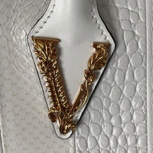 Puede incluir: Primer plano de un bolso blanco con un patr&oacute;n texturizado similar al de un reptil. El bolso presenta un emblema dorado con la letra "V". El asa es blanca y el bolso parece ser de cuero.