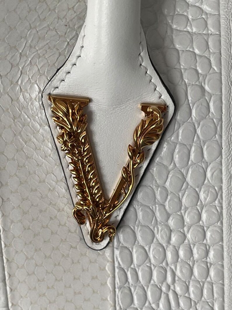 Puede incluir: Primer plano de un bolso blanco con un patr&oacute;n texturizado similar al de un reptil. El bolso presenta un emblema dorado con la letra "V". El asa es blanca y el bolso parece ser de cuero.