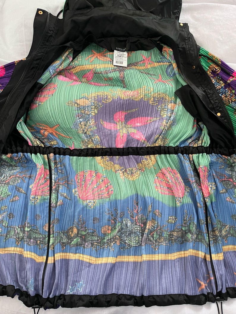 May include: A black hooded jacket with a vibrant, pleated interior lining featuring a colorful marine-themed print. The pattern displays pink starfish, seashells, and coral motifs against green and blue backgrounds, with a Versace brand label.