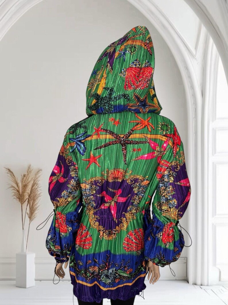 May include: A hooded jacket with a pleated texture, featuring a vibrant green base with a colorful print of red, orange, and blue starfish, seashells, and ornate gold-toned patterns. The jacket has long sleeves with adjustable drawstring cuffs.