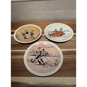 May include: Three vintage decorative plates with colorful illustrations. One plate depicts a couple dancing, another shows a car, and the third features figures with a dog. Each plate has a unique design and text.
