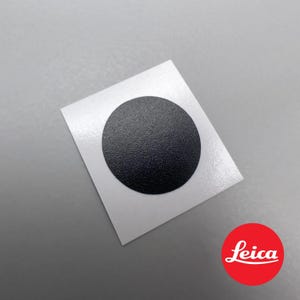 Pode incluir: Um adesivo circular preto com uma superfície texturizada sobre um fundo quadrado branco.