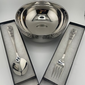 May include: A stainless steel serving bowl and serving utensils set. The bowl has the words "Bee Kind" and a bee graphic etched around the rim. A spoon and fork are displayed in individual black boxes.