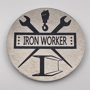 May include: A silver round metal pin with a black design. The design features a wrench and a hook, crossed over a steel beam. The text "IRON WORKER" is written in a banner above the beam.