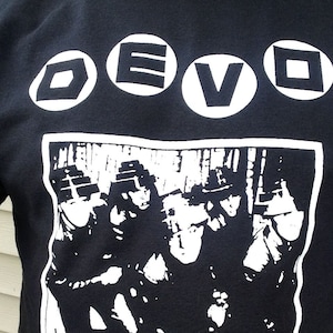 Puede incluir: Camiseta negra con un gráfico blanco de la banda DEVO. El gráfico incluye el nombre de la banda en círculos blancos y una imagen en blanco y negro de los miembros de la banda.