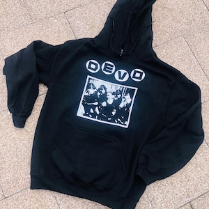 May include: Black hooded sweatshirt with a front pocket and drawstring hood. The sweatshirt features the band name "DEVO" in white block letters above a black and white graphic of the band members.