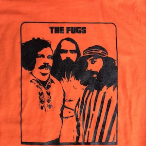 Op de afbeelding: Een zwart-wit afbeelding van drie mannen in een band genaamd "The Fugs". De afbeelding is op een oranje T-shirt.