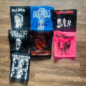 May include: A selection of vintage band t-shirts featuring iconic bands such as David Bowie, Black Sabbath, Thin Lizzy, Celtic Frost, Corrosion of Conformity, The Stooges, and The Smiths. Each shirt displays band names and graphics.