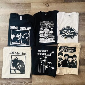 May include: Seven graphic t-shirts in black, white, and cream. The shirts display band logos and text, including "Radio Birdman," "Suicidal Tendencies," "Melvins," "Modest Mouse," "Buzzcocks," and "S&S Performance." The shirts are laid flat.