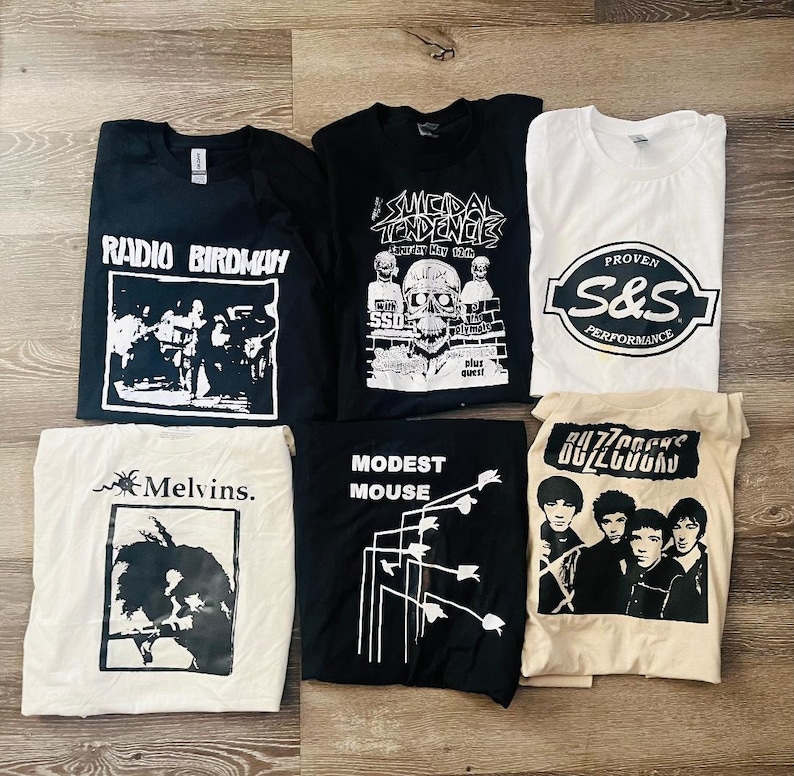May include: Seven graphic t-shirts in black, white, and cream. The shirts display band logos and text, including "Radio Birdman," "Suicidal Tendencies," "Melvins," "Modest Mouse," "Buzzcocks," and "S&S Performance." The shirts are laid flat.