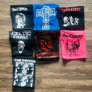 May include: A selection of folded band t-shirts featuring iconic rock and metal bands. Shirts include David Bowie, Black Sabbath, Thin Lizzy, Celtic Frost, Corrosion of Conformity, The Stooges, and The Smiths. The shirts are various colours, including black, blue, and pink.