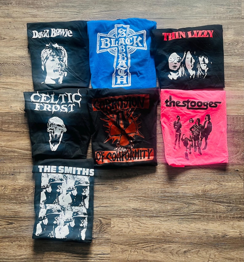 May include: A selection of folded band t-shirts featuring iconic rock and metal bands. Shirts include David Bowie, Black Sabbath, Thin Lizzy, Celtic Frost, Corrosion of Conformity, The Stooges, and The Smiths. The shirts are various colours, including black, blue, and pink.