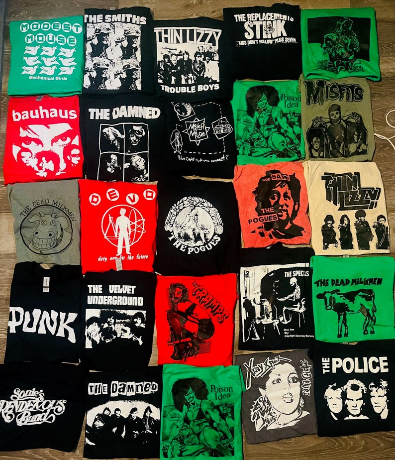 May include: A selection of band t-shirts in various colours, including black, red, green, and grey. The shirts feature band names and graphics such as "The Smiths," "Thin Lizzy," "The Damned," and "The Pogues." The shirts are laid out flat.