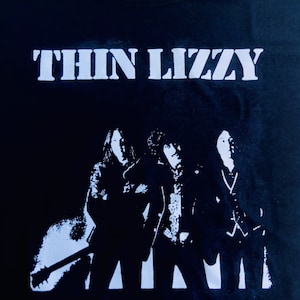 Può includere: Maglietta nera con una stampa bianca della band Thin Lizzy. La stampa presenta tre membri della band in silhouette.
