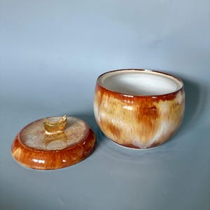 Könnte beinhalten: Ein Keramiktopf mit Deckel in Braun-, Beige- und Weißtönen. Der Topf hat eine glänzende Oberfläche, und der Deckel ist mit einem goldfarbenen Griff verziert. Der Innenraum ist weiß. Geeignet zur Aufbewahrung.