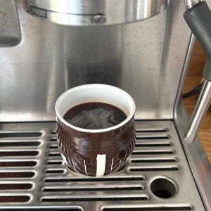 May include: A small, round, dark brown ceramic cup with a white interior and vertical white painted stripes, filled with black coffee. The cup sits on the stainless steel drip tray of an espresso machine, positioned directly beneath the group head.