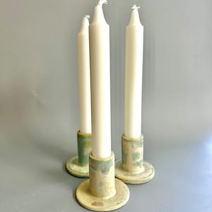 May include: Three white candles in a set of three ceramic candle holders. The holders are a light green and cream colour with a speckled glaze.