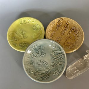May include: Three ceramic plates in shades of yellow-green, tan, and blue-green, each with a detailed dragon design. The plates are round with a slightly raised edge. A clear crystal is visible in the background.