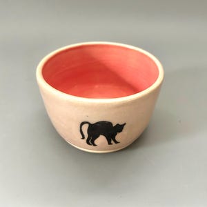 May include: A small ceramic bowl with a pink interior and a black silhouette of a cat on the exterior.